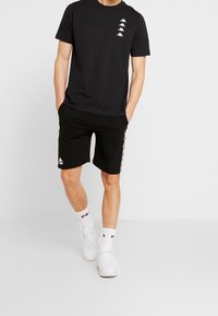 Černé bavlněné tričko s bílými potisky loga na hrudi, v kombinaci s černými šortkami s bočními pruhy a logem. Doplňuje bílé tenisky.