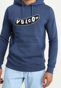 Sudadera de color azul marino hecha de una tela suave, con un bolsillo tipo canguro y un logotipo gráfico en blanco de "VOLCOM" en la parte frontal. Mangas largas y dobladillo acanalado.