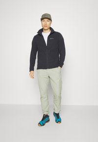 Schwarze Fleecejacke mit Reißverschluss und hohem Kragen, kombiniert mit hellgrünen Hosen und blauen Wanderschuhen. Enthält eine Kappe mit einem Logo.