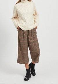 Pull à col roulé en tricot crème avec une texture en câble, associé à un pantalon cropped large à motif écossais brun et rouge et des bottines noires.