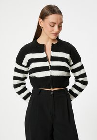 Mustavalkoraidallinen lyhyt cardigan, jossa on ribbitetty pinta, lyhyet hihat ja vetoketjukiinnitys. Puettu ylös korkeavyötäröisten mustien housujen päälle.