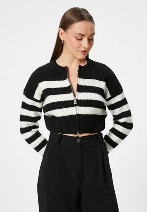Mustavalkoraidallinen lyhyt cardigan, jossa on ribbitetty pinta, lyhyet hihat ja vetoketjukiinnitys. Puettu ylös korkeavyötäröisten mustien housujen päälle.