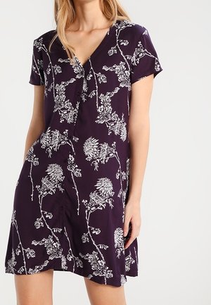 Woman wearing a short-sleeve, deep purple dress with white floral patterns and a V-neckline, standing against a plain light background.