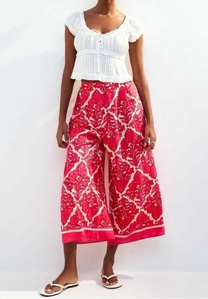 Personne portant un haut blanc à manches courtes froncées, un pantalon large court avec motif floral rouge, et des sandales blanches, debout devant un fond blanc.