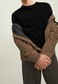 Pull noir à col rond et coupe ajustée, superposé sous une veste en suède marron avec doublure grise. Tissu gris visible aux manches ; design minimaliste.