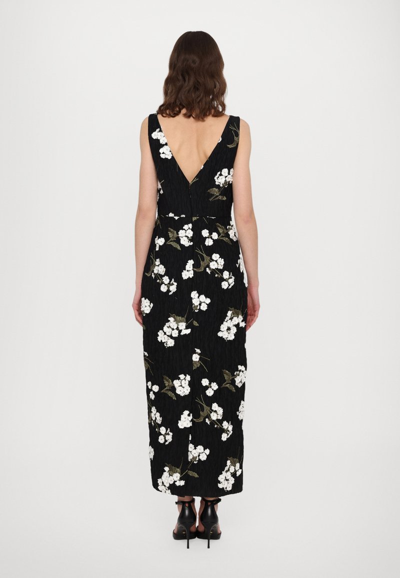 Robe maxi noire à fleurs avec un dos en V, ornée de broderies blanches et vertes sur un tissu froissé. Portée avec des sandales noires à talon et bride cheville.