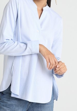 Frau trägt eine hellblaue Langarmbluse mit kragenlosem Ausschnitt und dunkelblaue Jeans, richtet mit den Händen einen Ärmel.