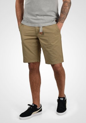 Shorts beige avec une coupe décontractée, dotés d'une taille élastique, d'un cordon tressé et de poches latérales. Associés à des baskets noires.