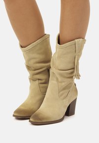 Botas de tobillo de ante beige con un diseño de caída, punteras puntiagudas y tacones apilados de 10 cm en marrón. Presentan cordones decorativos y cremalleras laterales.