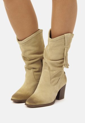 Bottes de cheville en daim beige avec un design froissé, bouts pointus et talons empilés marron de 10 cm. Présente des lacets décoratifs et des zips sur les côtés.