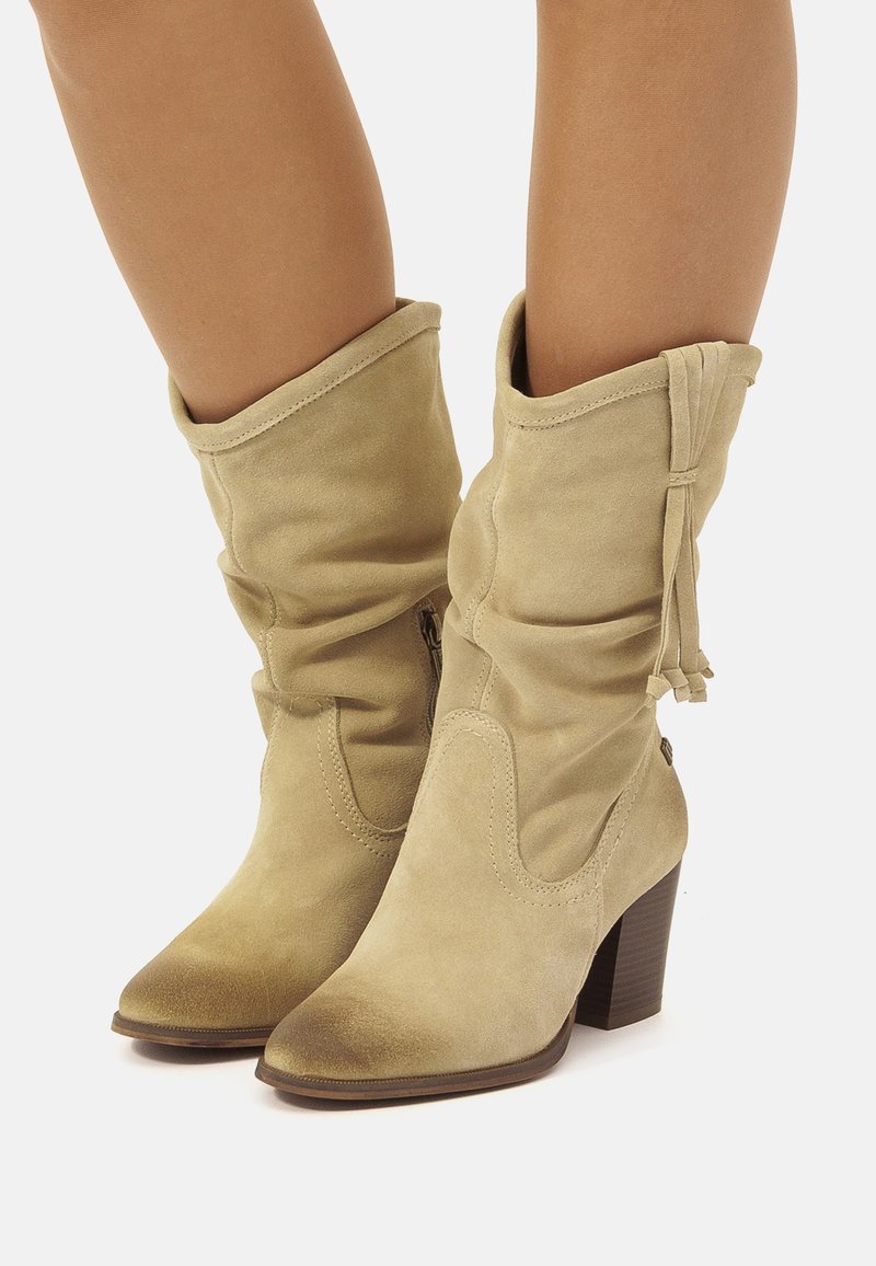 Botas de tobillo de ante beige con un diseño de caída, punteras puntiagudas y tacones apilados de 10 cm en marrón. Presentan cordones decorativos y cremalleras laterales.