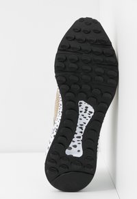 Semelle de chaussure de course avec des crampons circulaires noirs et une semelle intermédiaire blanche arborant un motif tacheté noir et beige.