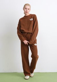 Brauner Fleece-Trainingsanzug mit langen Ärmeln und passenden Hosen. Mit einem weißen Logo auf der Brust und der Hüfte. Lockerer Schnitt und Kordelzugbund.