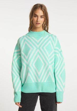 Frau trägt einen mintgrünen und weißen Pullover mit geometrischem Muster, mit gerippten Bündchen und Saum, kombiniert mit schwarzen Hosen, steht vor weißem Hintergrund.