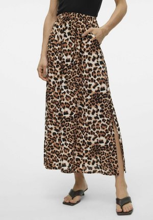 Frau trägt einen Midi-Rock mit Leopardenmuster und seitlichem Schlitz, ein schwarzes ärmelloses Oberteil und schwarze Sandalen mit Absatz, steht mit einer Hand in der Tasche.