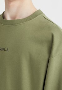 Oliwkowa bluza wykonana z miękkiego materiału, z prążkowanym dekoltem i prostym czarnym logo z przodu. Gładka struktura i luźny krój.