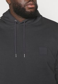 Hoodie gris foncé en tissu doux, avec une poche kangourou, des cordons de serrage aux embouts argentés et un petit patch carré.