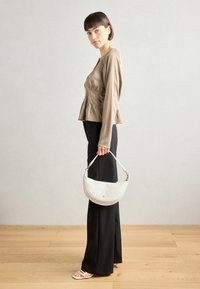 Haut en satin beige à manches longues avec un design plissé, associé à un pantalon noir taille haute. Le mannequin tient un sac à main texturé blanc.