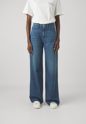 Personne portant un jean bleu taille haute à jambes larges, associé à un t-shirt blanc et des baskets blanches, debout devant un fond uni.