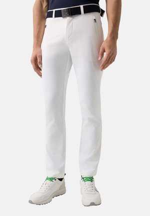 Muž nosící bílé úzké kalhoty, černý pásek a bílé tenisky se zelenými tkaničkami, stojící s uvolněnýma rukama po stranách.