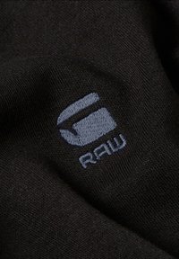 Schwarzer Stoff mit einer weichen Textur, versehen mit einem gestickten Logo in Hellblau, das "RAW" liest. Einfache, minimalistische Designsprache.