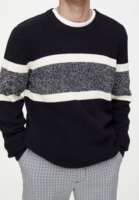 Pull en tricot bleu marine, avec de larges rayures horizontales blanches et grises. Texture côtelée, col rond. Associé à un pantalon à carreaux.