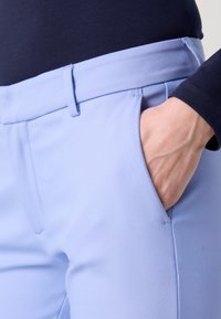 Gaiši zili bikses ar gludu tekstūru, ar taisnu fronti, sānu kabatām un pieguļošu dizainu. Jostasvieta ir nedaudz pacelta.