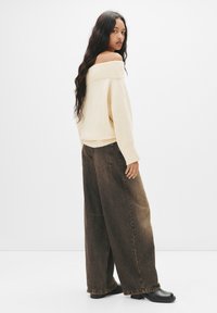 Maglione crema off-the-shoulder con polsini a costine abbinato a jeans a gamba larga marrone scuro, con tasche posteriori e vestibilità rilassata. Stivaletti neri alla caviglia.