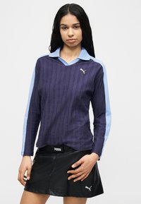 Γυναίκα με μακριά σκούρα μαλλιά, φορώντας μωβ και γαλάζιο πουκάμισο με μακριά μανίκια και μαύρη  φούστα Puma, έμπροσθεν απλού φόντου.