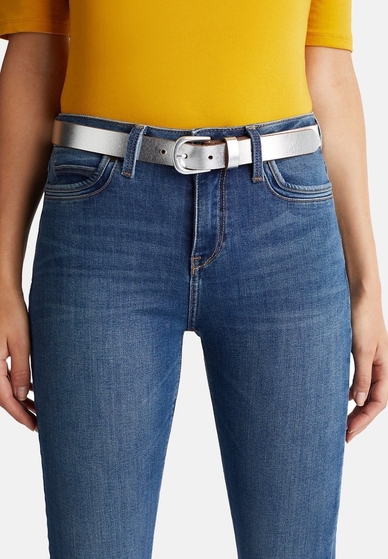 Personne portant un jean en denim bleu avec une ceinture métallique argentée et une chemise jaune, montrée de la taille jusqu’au milieu de la cuisse.