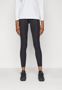 Leggings deportivos negros con un diseño ajustado, textura suave, cintura de altura media y detalle del logo a lo largo del lado izquierdo; combinados con zapatillas negras.