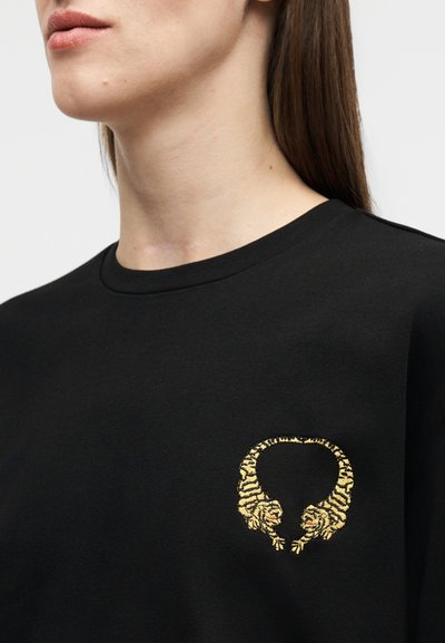 Κοντινό πλάνο ενός ατόμου που φοράει μαύρο πουκάμισο με κεντητό σχέδιο δύο κίτρινων τίγρεων που κοιτάζονται μεταξύ τους σχηματίζοντας έναν κύκλο.