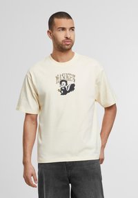 Lysbeige kortærmet t-shirt med et grafisk motiv af en sort tegneserie-skunk og ordet "MARKET" ovenfor. Blødt bomuldsstof, løs pasform.