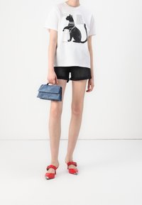 T-shirt bianca in cotone con grafica di un gatto nero, abbinata a pantaloni shorts in denim scuro e scarpe in pelle lucida rossa con punta affilata e fibbia.