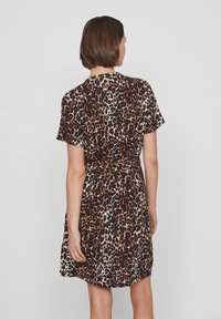 Žena s kratkom smeškom koja nosi haljinu na koljenima kratkih rukava s uzorkom leopard tiska, okrenuta leđima prema jednostavnoj svjetloj pozadini.