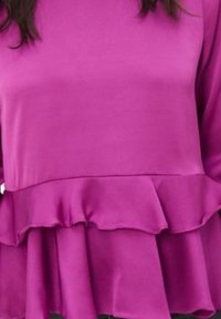 Blusa morada hecha de una tela suave con mangas largas y un dobladillo con volantes. El diseño presenta un estilo peplum escalonado para añadir textura.