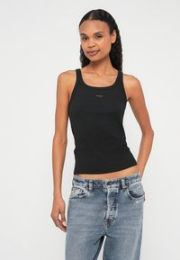 Μαύρο εφαρμοστό τοπ με φαρδιές τιράντες, τετράγωνο ντεκολτέ και μικρή λεπτομέρεια λογότυπου στο στήθος, σε συνδυασμό με φθαρμένο ανοιχτό μπλε τζιν.