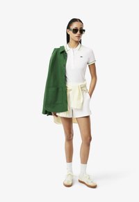 Polo bianco con rifiniture verdi e gialle, abbinato a pantaloni corti bianchi. Giacca verde gettata sulle spalle, scarpe casual e occhiali da sole inclusi.