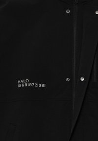 Schwarze Jacke vorne mit silbernem Reißverschluss und Druckknöpfen, mit weißem aufgedrucktem Text "HALO 196819721981" auf der linken Seite.