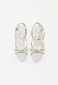 Silver strappy high-heeled sandals with a smooth white sole, decorative knot detail, and ankle straps featuring metallic accents.