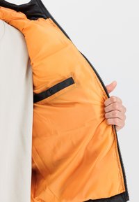 Melnā ārējā jaka, oranža oderējuma, ar rāvējslēdzēju priekšā, iekšējā kabata ar melnu akcentu. Gluda tekstūra, viegls dizains, ikdienišķs stils.