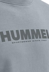 Felpa azzurra chiara con grande scritta in grigio scuro "HUMMEL" e "SPORTSWEAR SINCE 1923" in un carattere più piccolo. Tessuto morbido e текстurizzato.