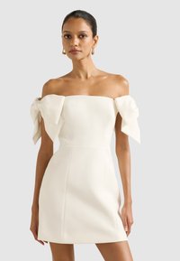 Bjela haljina bez naramenica s velikim mašnama, tijesno krojena na gornjem dijelu i A-linijom suknje. Glatka tekstura i minimalistički dizajn. Elegantna silueta.