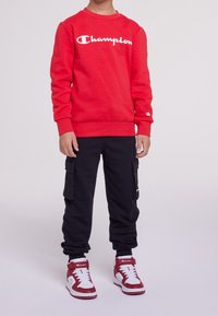 Felpa rossa in cotone con logo bianco "Champion", abbinata a pantaloni cargo neri con tasche laterali e sneakers alte rosse e bianche.