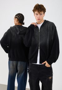 Černá mikina na zip s lehkým vyblednutím na ramenou, žebrovanými manžetami a klokaními kapsami; nošena dvěma modely s různými účesy.