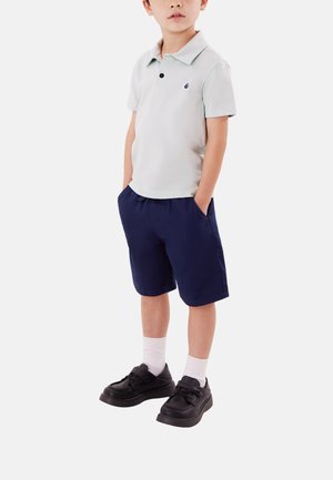Ragazzo giovane in piedi con le mani nelle tasche, indossa una polo grigio chiaro, pantaloncini blu navy, calzini bianchi e scarpe nere.