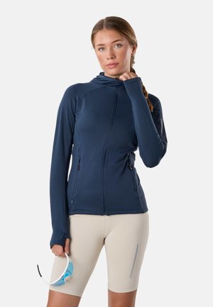 Kobieta z plecionymi włosami nosi granatową bluzę na zamek i beżowe krótkie spodenki kolarskie, trzymając w jednej ręce białe i niebieskie sportowe okulary przeciwsłoneczne.