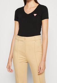 Zwarte V-hals T-shirt met korte mouwen, voorzien van een rood en wit logopatch. Gecombineerd met lichtbeige, high-waisted op maat gemaakte broek.