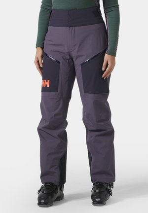 Person iført lilla og sorte skibukser med orange logo, sorte skistøvler og grøn langærmet top, stående mod hvid baggrund.