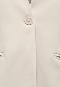 Hellbeiger Blazer mit strukturierter Stoffqualität, ausgestattet mit einem einzelnen runden Knopfverschluss und zwei diagonalen Taschen mit kontrastierender Einfassung.